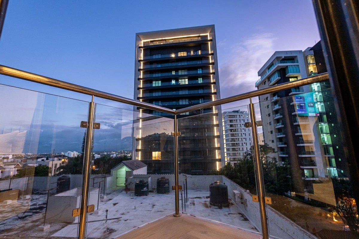 A private balcony offers a view of neighboring buildings illuminated at dusk. Glass railings enhance the open feel, while the city skyline creates a vibrant backdrop against the twilight sky. The tranquil atmosphere is perfect for enjoying peaceful moments.