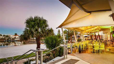 An outdoor dining area features tables and chairs under a large canopy, surrounded by palm trees and lush greenery. The setting is illuminated softly as sunset colors create a serene atmosphere along the waterfront.