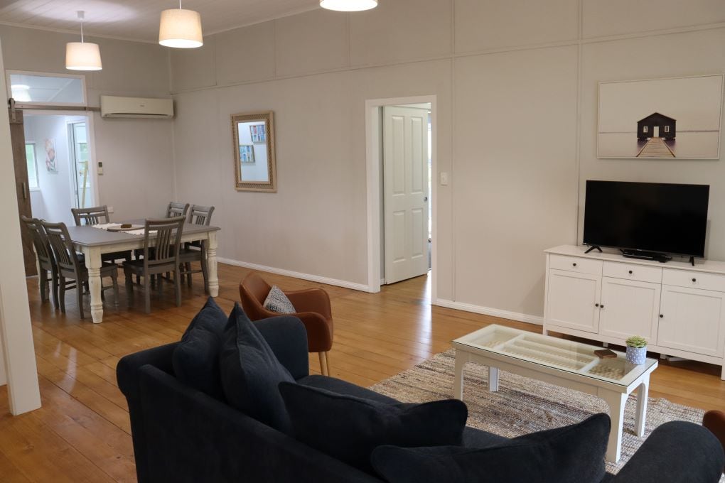 A well-lit living area features a comfortable dark sofa with accent chairs, a coffee table, and a television on a white cabinet. Natural wood flooring complements the room. A dining area is visible in the background, showcasing a table with multiple chairs.