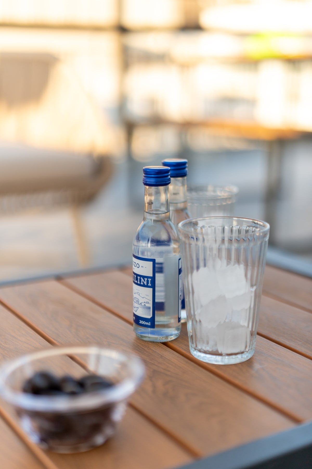 A table is set outdoors, featuring two small bottles labeled ‘Bellini’ alongside a tall glass filled with ice. A bowl containing dark olives is also present, providing a casual and inviting setting for refreshments.