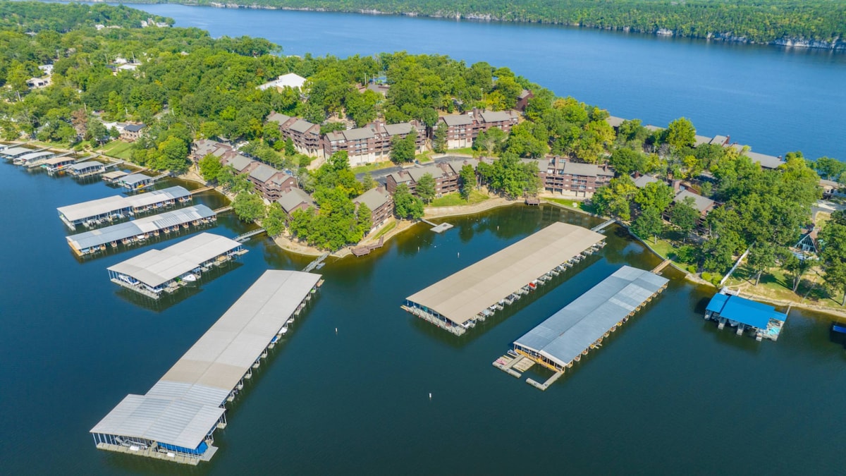 An aerial view captures the serene Knolls community, highlighting its lush greenery and well-maintained buildings. Docks extend into the calm waters, providing ample boat slips. The expansive lake stretches into the distance, framed by trees along the shoreline.