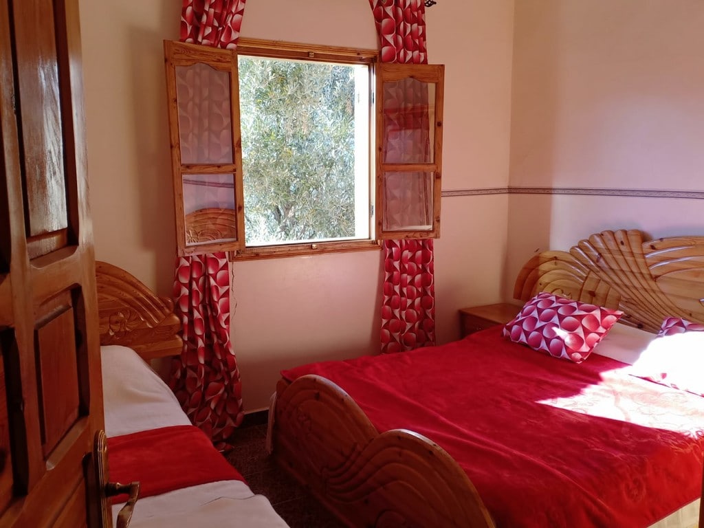 A cozy bedroom features two wooden beds with red bed linens and decorative pillows. Light streams in through a large window adorned with vibrant curtains, offering a view of greenery outside. The room is complemented by wooden furniture and a warm ambiance.