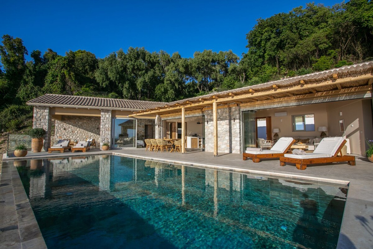 An inviting exterior view of Villa Parga showcases a private infinity pool reflecting the clear sky. Comfortable loungers are arranged poolside, while the spacious open-plan living area features large glass doors that connect the indoor and outdoor spaces, surrounded by lush greenery.