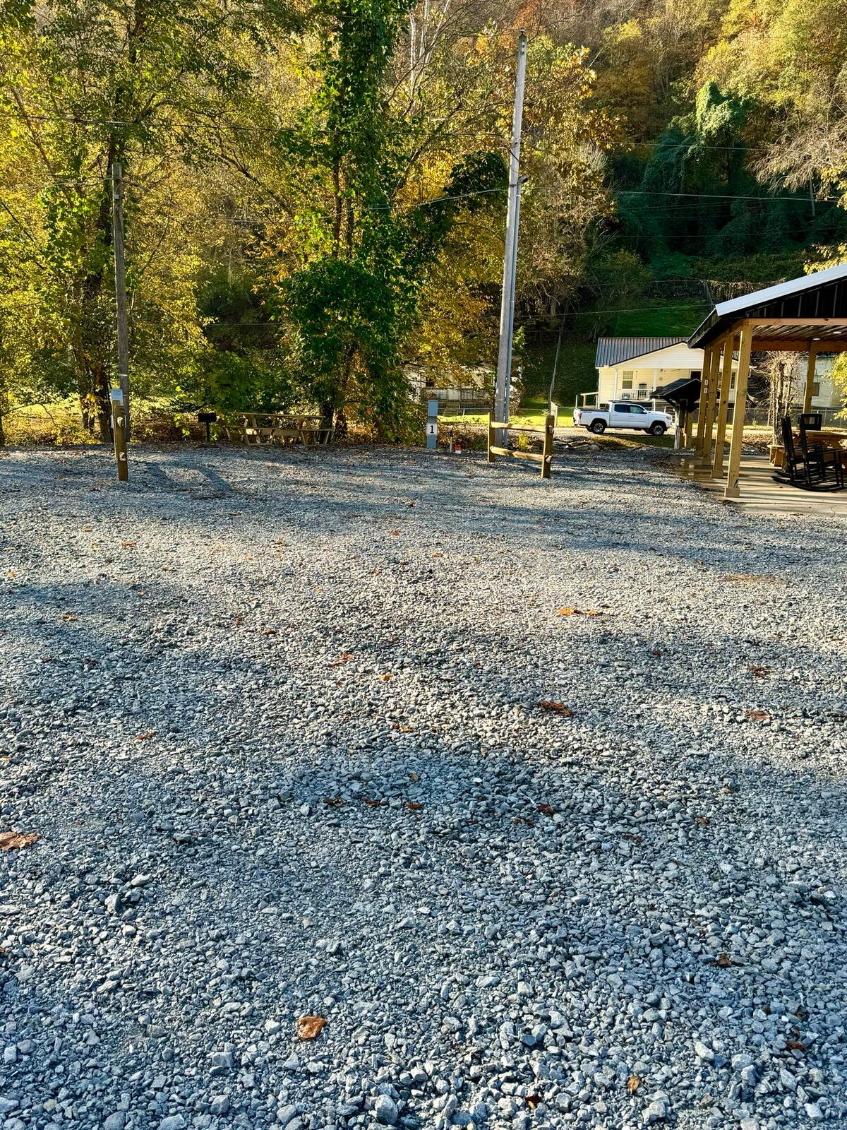 A gravel area is framed by trees in varying shades of green and yellow, indicative of the season. In the background, a vehicle is parked near a building, with utility poles visible along the roadway.