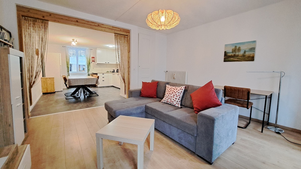A comfortable living area is highlighted by a grey sectional sofa adorned with red decorative pillows. A light wooden coffee table is positioned in front. The space opens to a dining area visible in the background, featuring a light fixture and a wooden table with chairs.