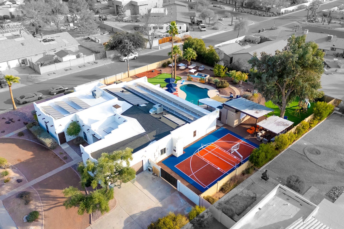 An aerial view showcases a spacious rental property featuring vibrant outdoor amenities. A private heated pool is visible alongside a hot tub, sportscourt, and kid's playground. Colorful game areas like a putting green and corn hole enhance the yard's lively atmosphere amid lush landscaping.