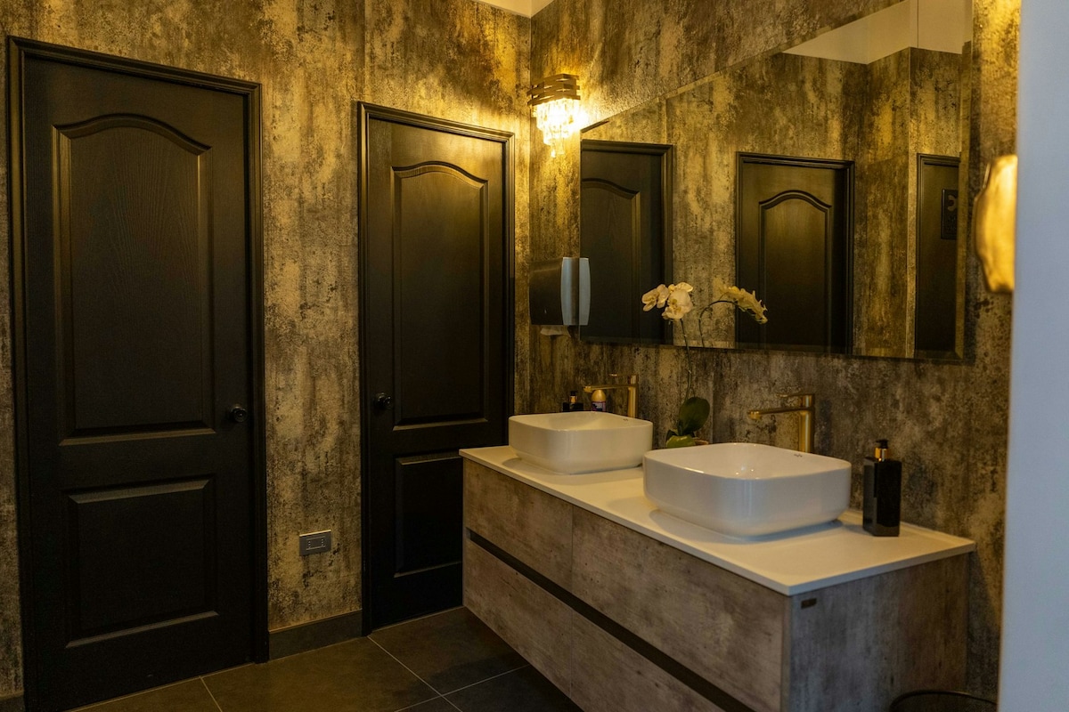 A contemporary bathroom features dual sinks set on a stylish countertop. Dark wood doors and rich textured walls complement the modern fixtures. Three mirrors reflect natural light, enhancing the spaciousness. Elegant lighting fixtures add a touch of sophistication to the overall design.
