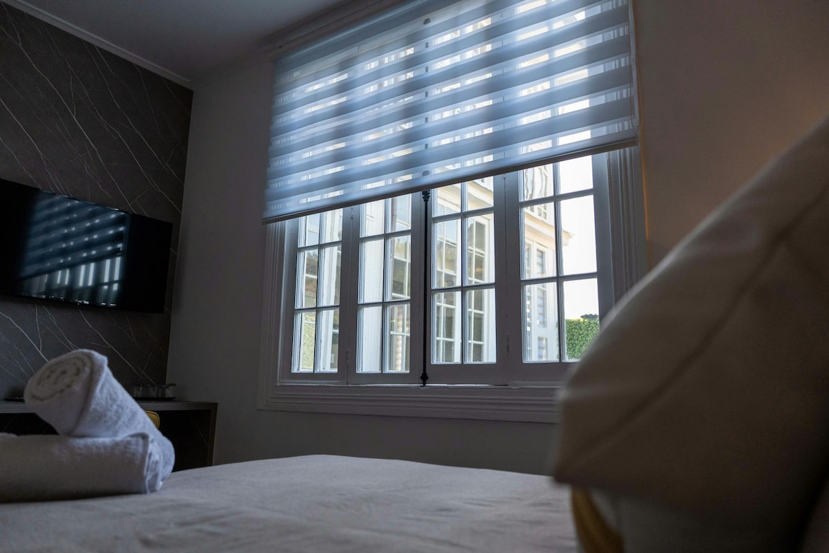 A spacious room is shown from a low angle, featuring a neatly made bed with rolled towels. Large windows are covered with sheer blinds, allowing soft light to filter in. A television is mounted on the wall opposite the window, reflecting the serene ambiance.