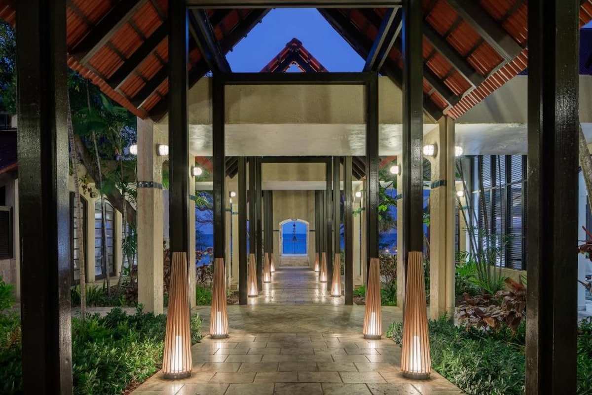 An elegant walkway is framed by tall columns and a vaulted wooden ceiling. Soft lighting highlights the pathway, guiding the eye toward an inviting entrance that opens up to lush greenery and the sea beyond.