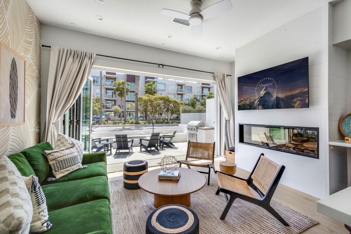 An open concept living space features a green sectional sofa and black accent chairs positioned around a round coffee table. A wall-mounted smart TV is accompanied by a modern fireplace underneath. Bifold doors lead to the outdoor patio, enhancing the connection to the outdoor area.