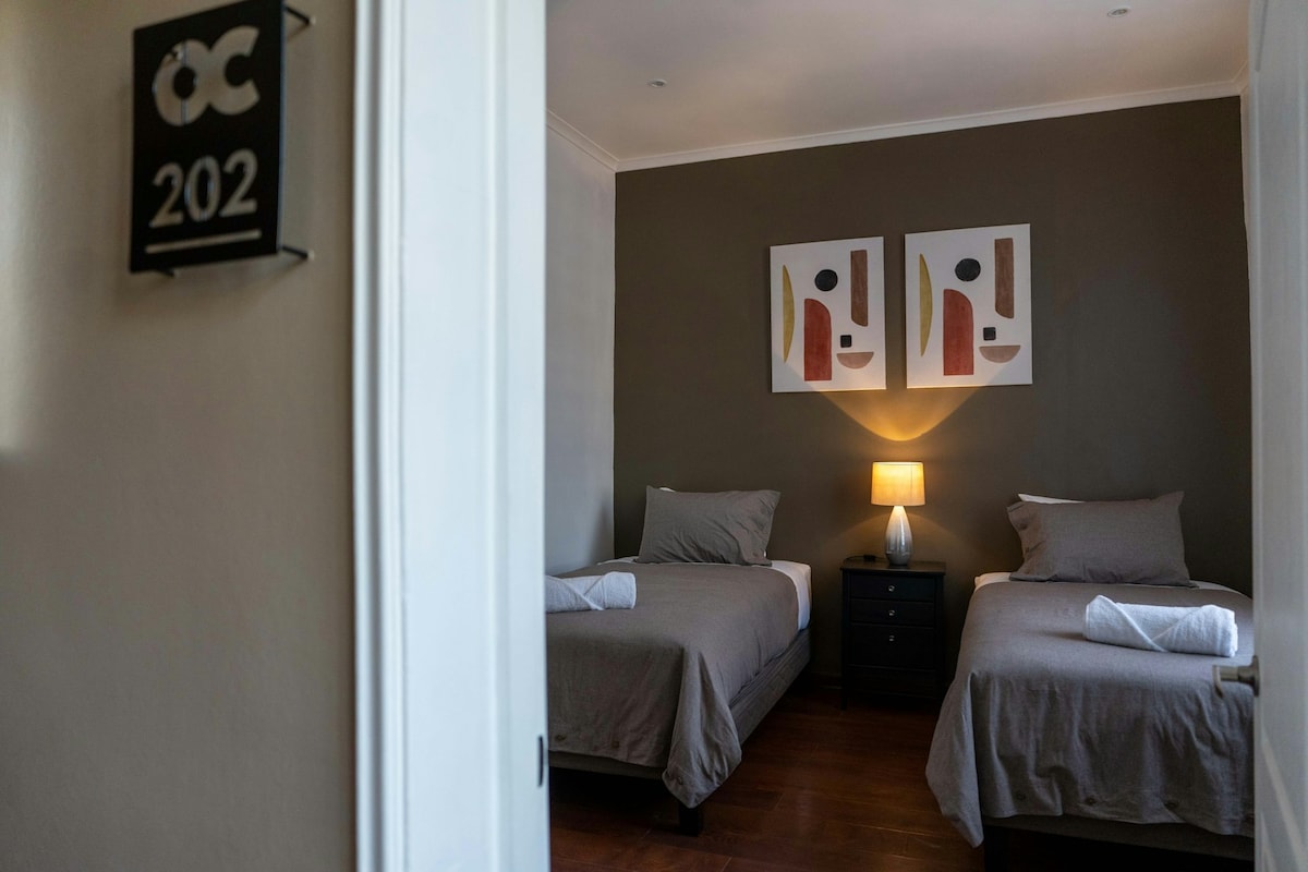 A guest room is featured with two spacious beds dressed in neutral linens. A nightstand holds a small lamp, providing practical lighting. Abstract artwork is displayed on the wall, and the room is complemented by a warm color palette, enhancing the overall atmosphere.