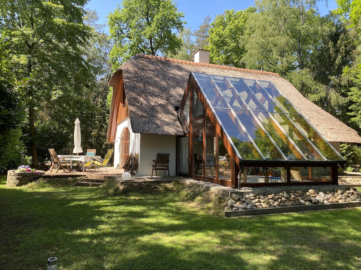 A holiday home is set amidst greenery, featuring a thatched roof and large glass panels that enhance natural light. Outdoor seating is provided on a wooden deck, accompanied by a sun umbrella. A landscaped garden area with stones adds to the charm of the setting.
