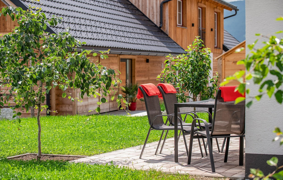 A patio area features a table surrounded by four chairs, with bright red blankets draped across the seats. Lush green grass and young trees enhance the outdoor setting, framed by wooden buildings in the background.