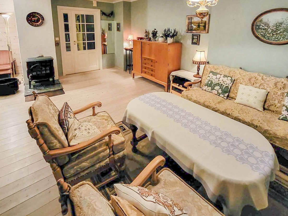 A spacious living room is presented, featuring a comfortable floral sofa and two matching armchairs. A wooden coffee table with a decorative tablecloth occupies the center. Soft lighting emanates from a chandelier and lamps on side tables, creating a cozy ambiance. A wood-burning stove adds warmth to the corner.