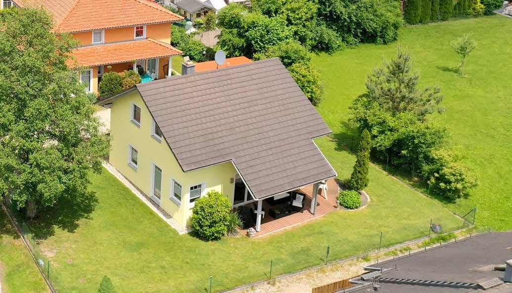 The image shows a spacious yellow house with a sloped roof, surrounded by a well-maintained green lawn. A patio area is visible beneath the porch, offering outdoor seating. The neighboring houses and trees frame the scene, enhancing the sense of privacy in the environment.