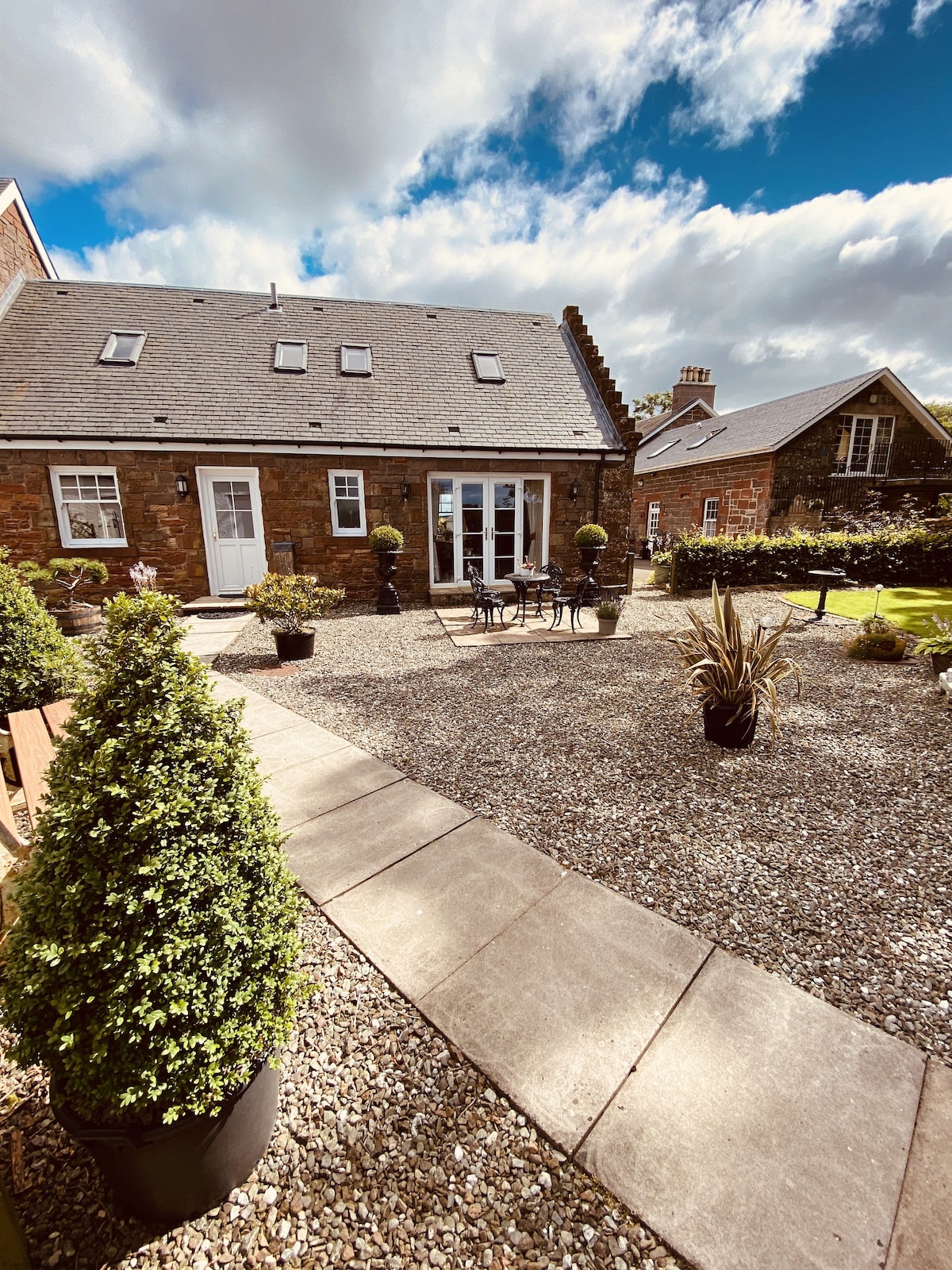 An outdoor area features a gravel path leading towards a two-story building with a sloped roof. Several potted plants and trees are positioned around a small seating arrangement with a table and chairs. Sunshine streams over the well-maintained garden space.