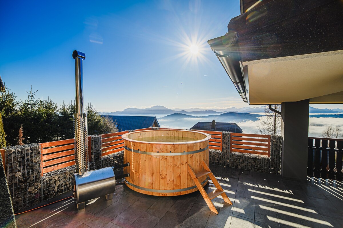 A private hot tub crafted from wood is positioned on a terrace, overlooking sweeping mountain views. Sunlight reflects off the water's surface, and a chimney is visible beside the tub. The surrounding area features wooden privacy panels and a paved floor, enhancing the outdoor experience.