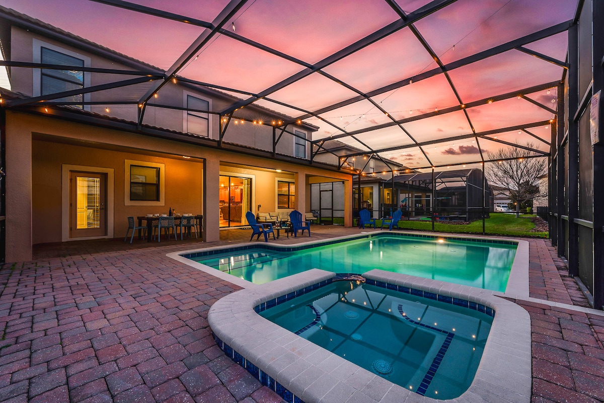 The outdoor area features a private pool with a hot tub, both enclosed under a screened lanai. A spacious patio is equipped with seating and a dining table, surrounded by twinkling lights. The sunset sky above adds a warm glow to the scene.