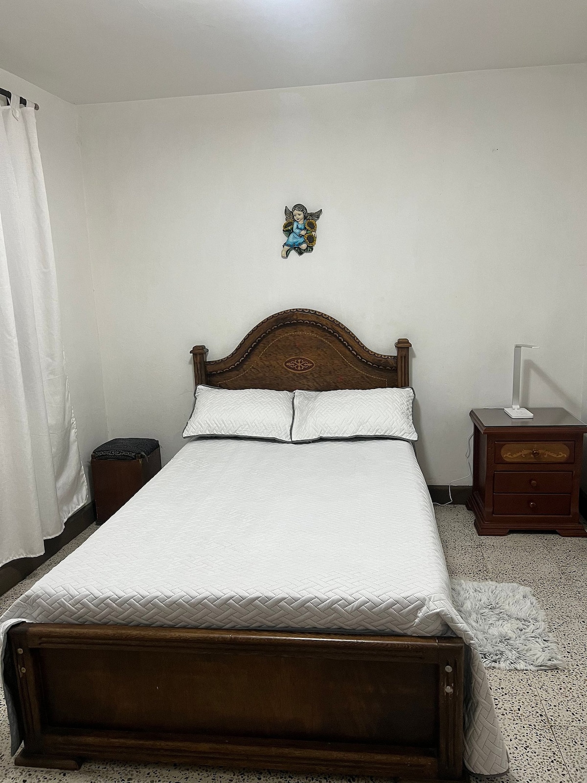 A cozy bedroom features a wooden bed with a simple mattress, adorned with white linens. A small bedside table with a lamp stands beside the bed. Natural light brightens the room through white curtains, while a decorative wall piece adds a personal touch.