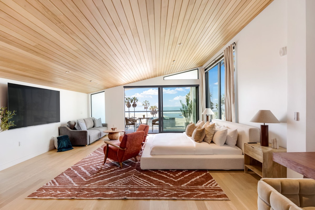 An open-concept living room features floor-to-ceiling glass doors that provide stunning views of the ocean. The space is furnished with a plush sectional, a cozy armchair, and a stylish coffee table, complemented by a warm wooden ceiling and a patterned area rug.