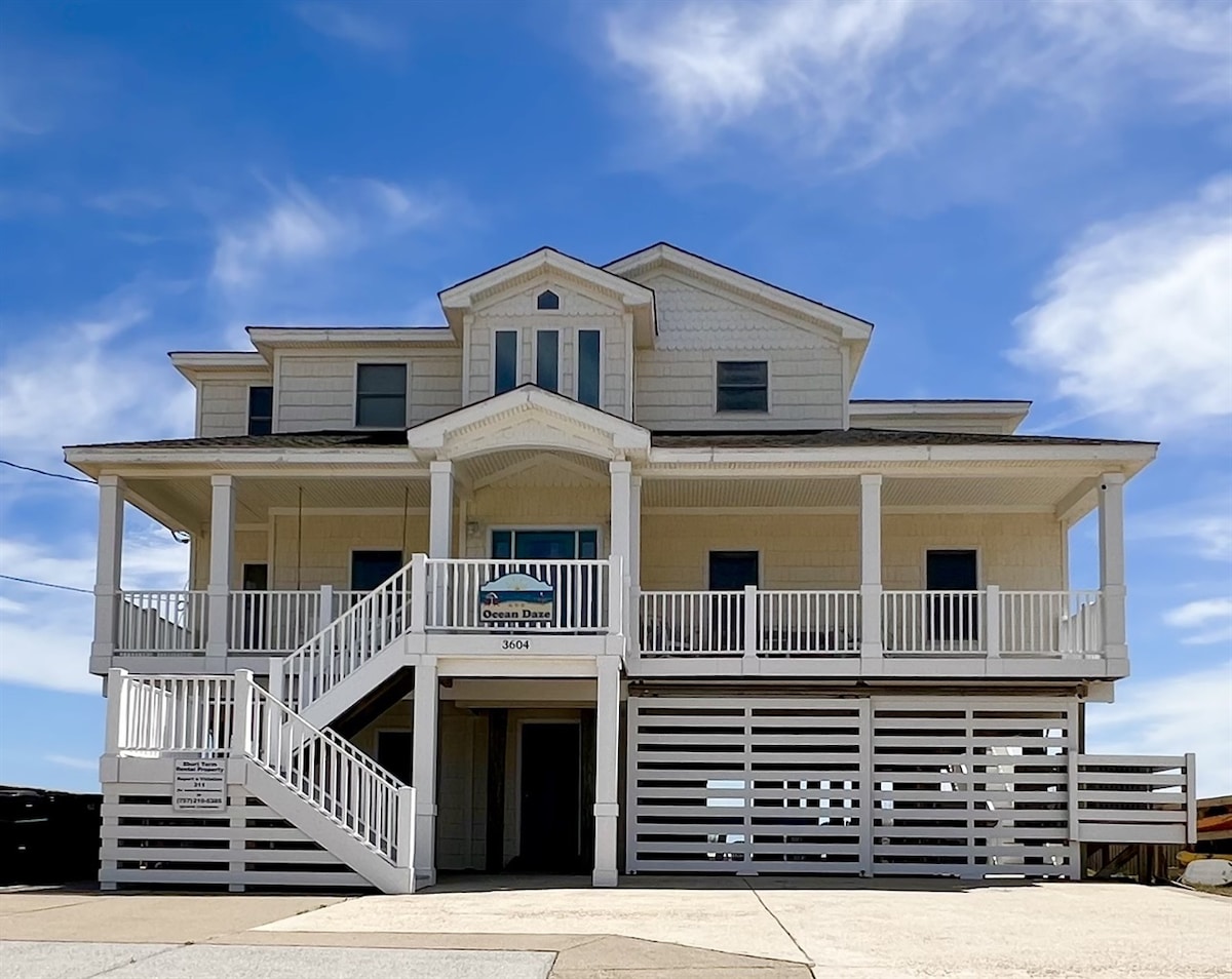 The exterior of the property showcases a three-story beach house, featuring a light-colored facade and multiple balconies. A wide staircase leads to the main entrance, complemented by a broad deck area beneath. Clear skies provide a bright backdrop, enhancing the coastal charm of the home.