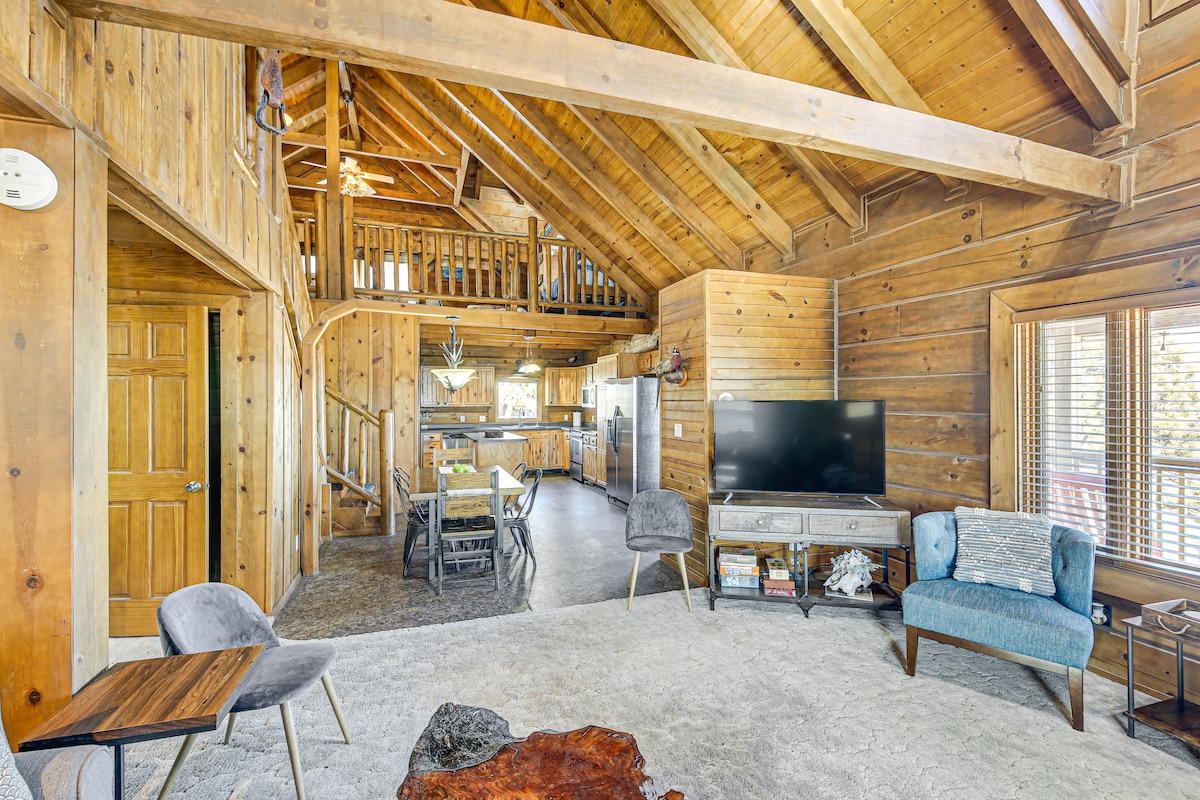 The living area features natural wood paneling, accentuated by high vaulted ceilings. A comfortable seating arrangement includes two chairs and a coffee table, with a large television mounted on a media console. A staircase leads to an upper loft area, visible in the background.