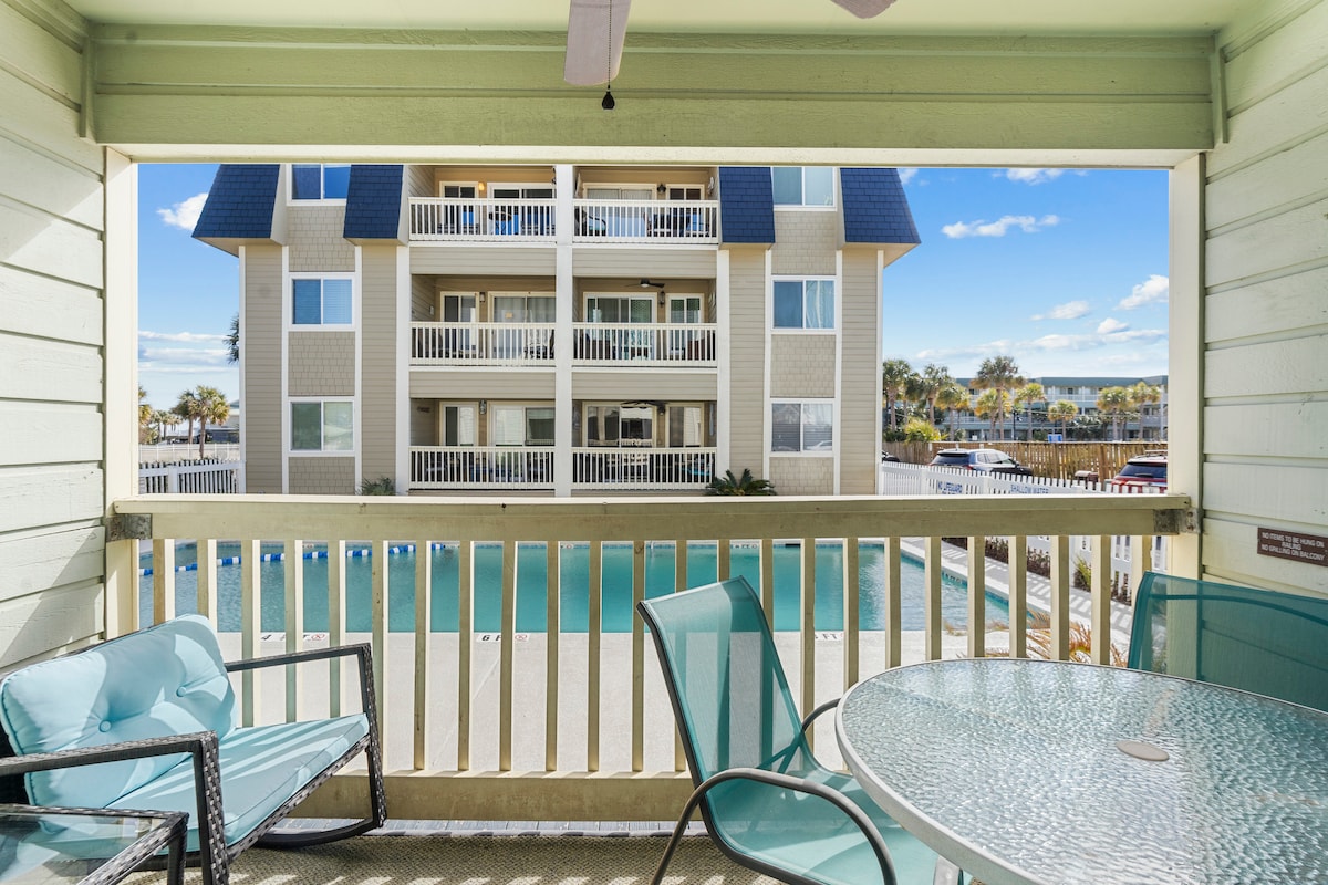 A private balcony features seating for six, including a glass table and lounge chairs. A view of the community pool and surrounding buildings is visible, highlighting the outdoor space and inviting atmosphere.