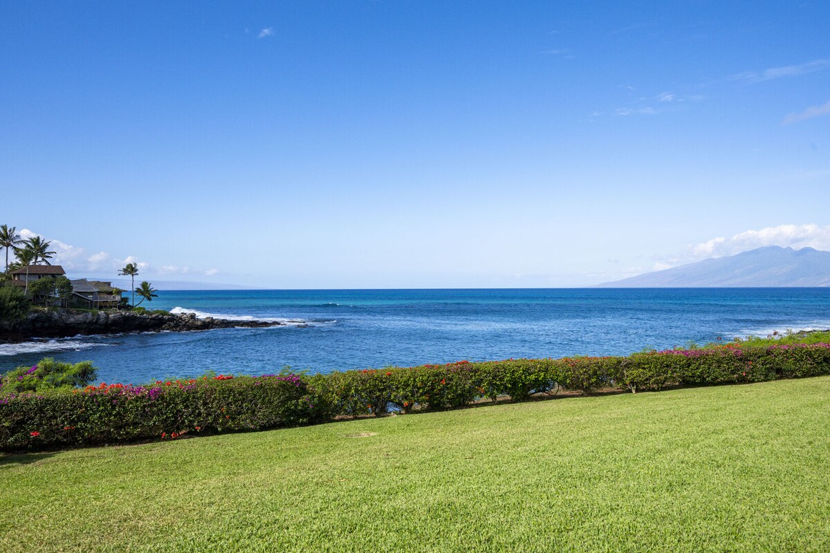 A vibrant view of the ocean is captured, showcasing clear blue waters stretching towards the horizon. Lush green grass leads to the shoreline, framed by a colorful border of blooming flowers and palm trees swaying gently in the breeze.
