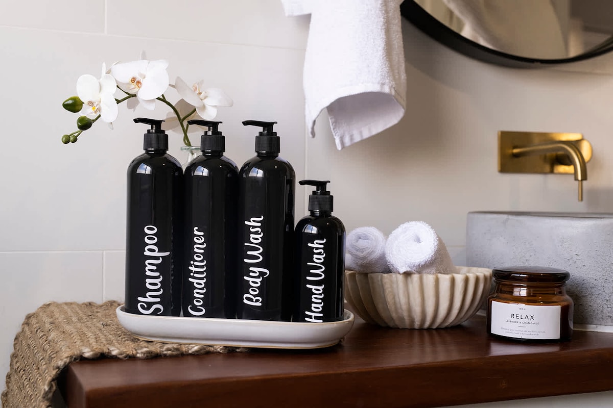 A neatly arranged bathroom counter is displayed, featuring black bottles labeled for shampoo, conditioner, body wash, and hand wash. A small white dish holds rolled towels beside a decorative candle labeled 'Relax'. Soft textures and organic shapes create an inviting atmosphere.