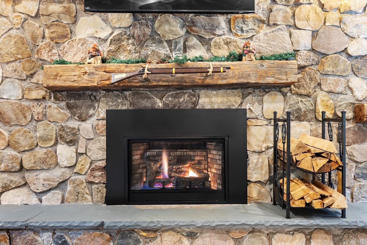 A stone fireplace is framed by a rugged stone wall, featuring a wood mantel adorned with decorative items. A metal log holder stands beside neatly stacked firewood, while a warm glow emanates from the fire, inviting relaxation and comfort.