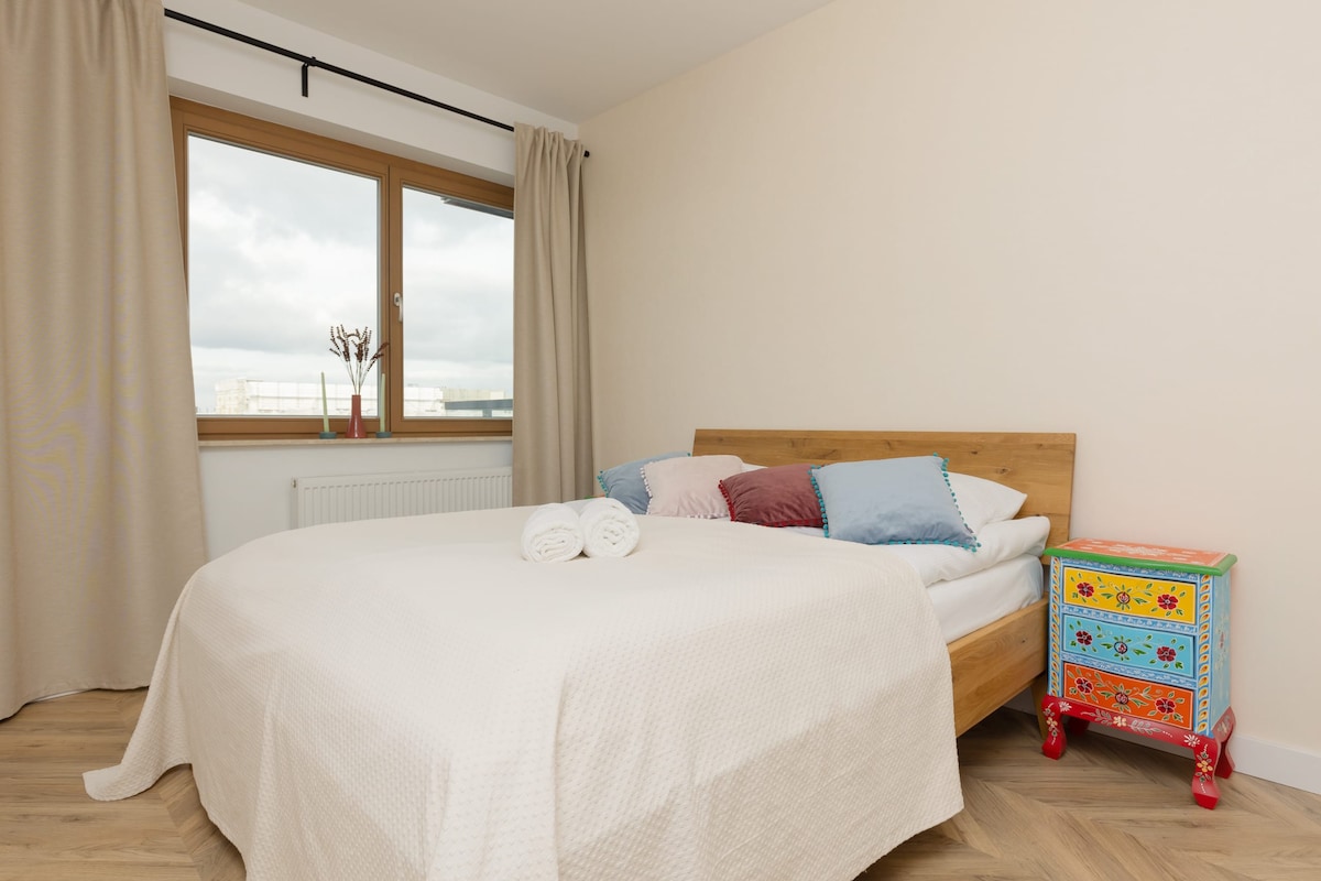 غرفة نوم تحتوي على سرير أبيض وأرضية خشبية ووحدة تخزين ملونة صغيرة لألعاب الأطفال. نافذة كبيرة مع ستائر تسمح بدخول أشعة الشمس.
