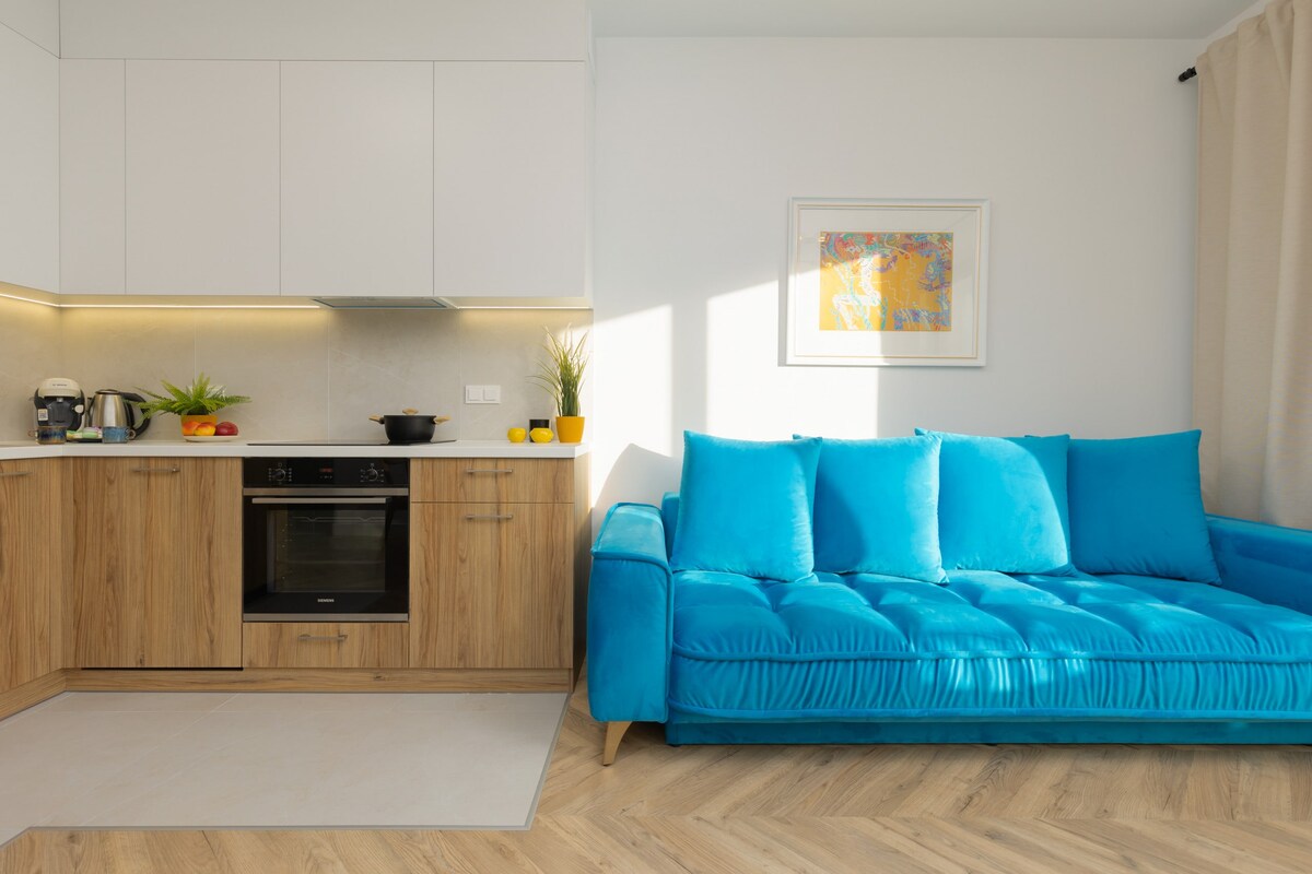 غرفة معيشة حديثة مع أريكة زرقاء زاهية وأرضية خشبية ومطبخ مفتوح مع خزانات خشبية خفيفة. المساحة مضاءة جيدًا بالضوء الطبيعي.
