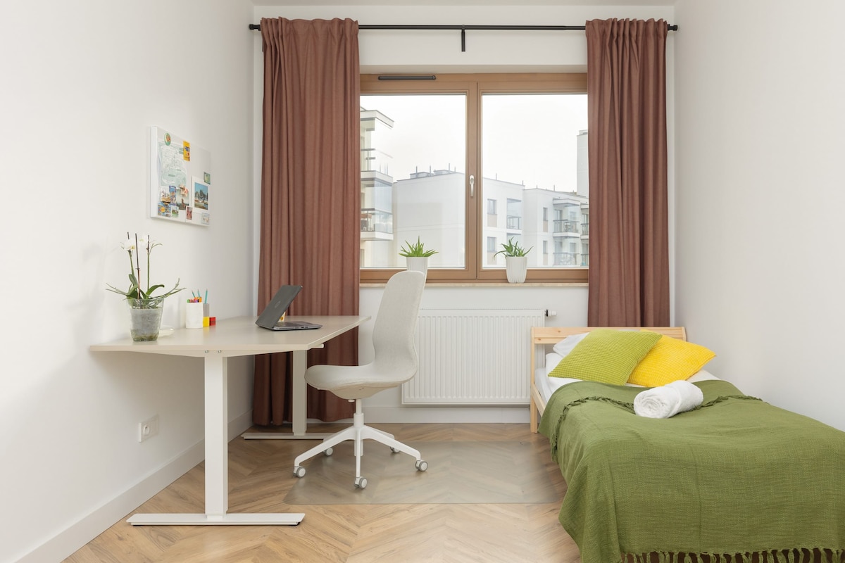 زاوية مكتب منزلية صغيرة مع مكتب أبيض وكرسي وسرير أخضر في مكان قريب. توفر النافذة إطلالة على أفق المدينة من بعيد.