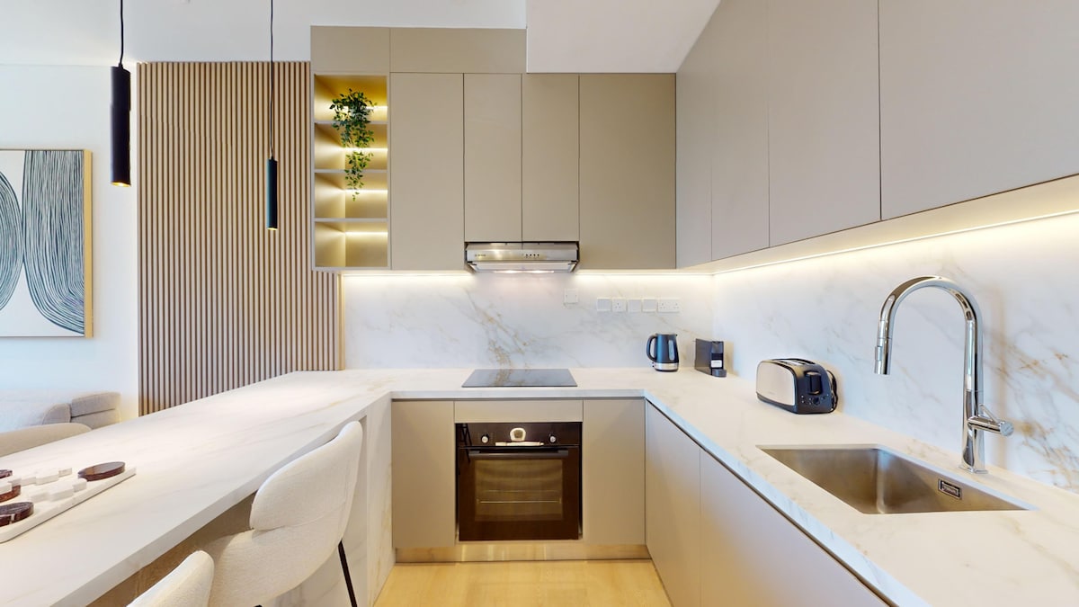 A modern, fully equipped open-plan kitchen features sleek cabinetry and a polished marble countertop. Kitchen appliances, including a toaster, kettle, and coffee machine, are visible. Stylish bar stools are arranged at the counter, and subtle lighting enhances the functional space.