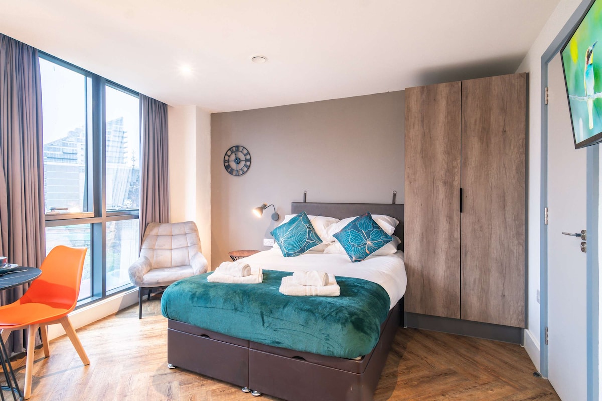 A modern studio bedroom features a plush double bed covered in a deep green throw and crisp white linens. Natural light enters through spacious windows, highlighting stylish furnishings. An armchair and dining table accompany a wall-mounted TV, with wooden flooring adding warmth to the contemporary design.