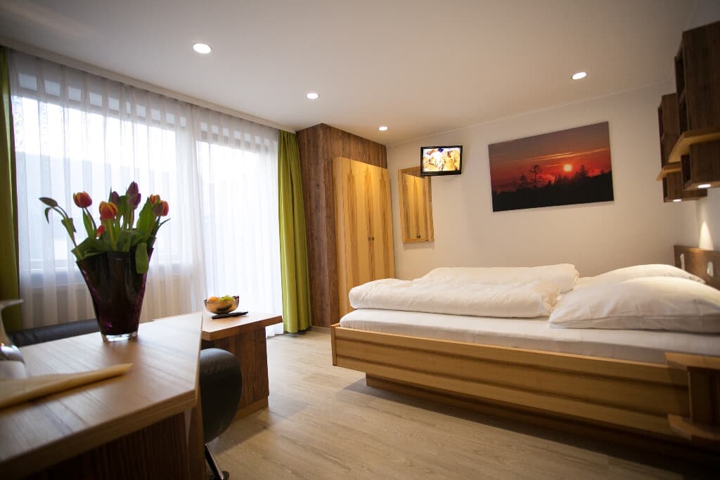 A well-lit room features a spacious bed with white bedding against a backdrop of natural wood accents. A flat-screen TV is mounted on the wall, and a bouquet of tulips sits on a wooden table. Soft lighting enhances the overall ambiance, complemented by large windows with sheer curtains.