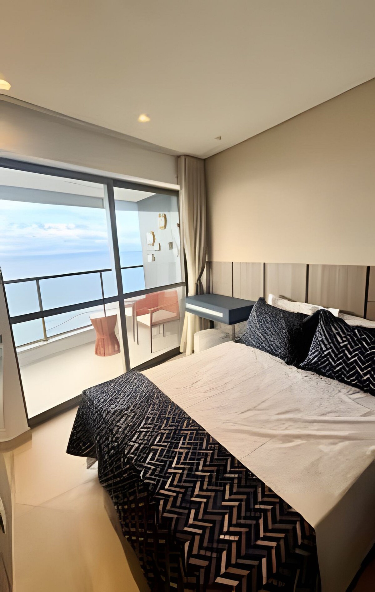 A cozy bedroom is presented with a spacious bed dressed in a navy and white patterned blanket. Floor-to-ceiling glass doors open to a balcony, offering views of the ocean. A small desk and two chairs are positioned nearby, completing the inviting atmosphere.