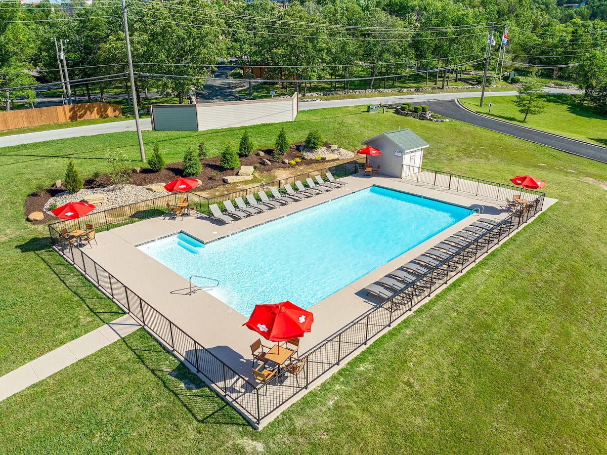 An aerial view of a shared outdoor pool is presented, surrounded by a green lawn. Red umbrellas provide shade for several lounge chairs, while additional seating is arranged near the poolside. The fenced area adds a layer of privacy to this inviting recreational space.