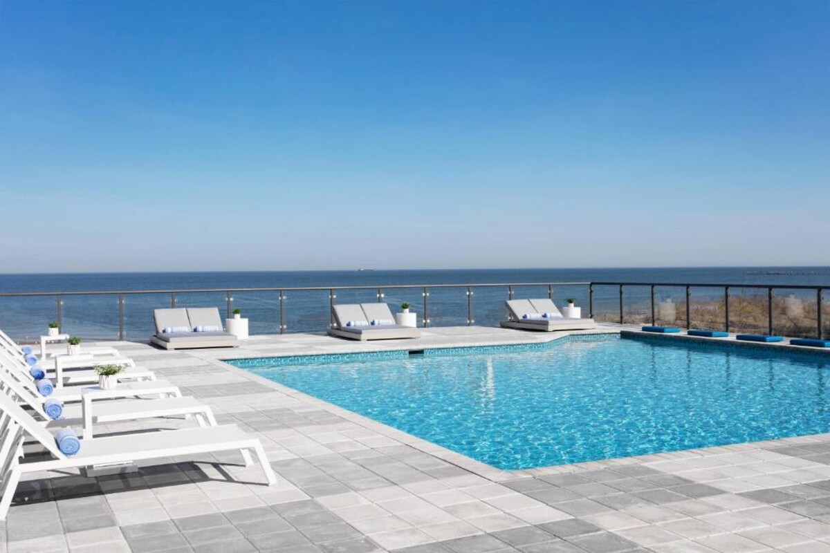An outdoor pool is framed by a spacious deck lined with white loungers, each adorned with rolled blue towels. Beyond the pool, expansive views of the tranquil water stretch towards the horizon under a clear blue sky.