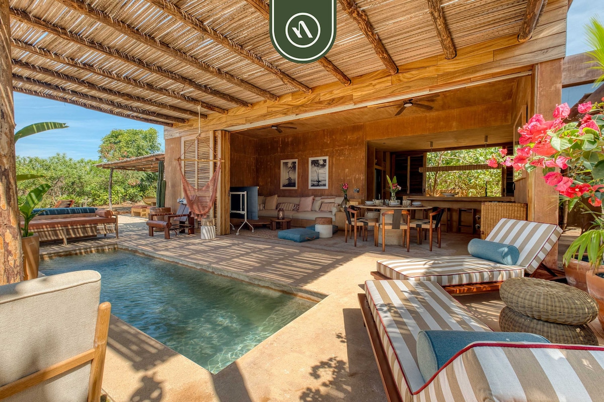 An inviting outdoor space features a private pool surrounded by wooden lounge chairs with striped cushions. A dining table under a thatched roof is visible, accompanied by comfortable seating arrangements and decorative plants, creating a relaxed atmosphere for outdoor living.
