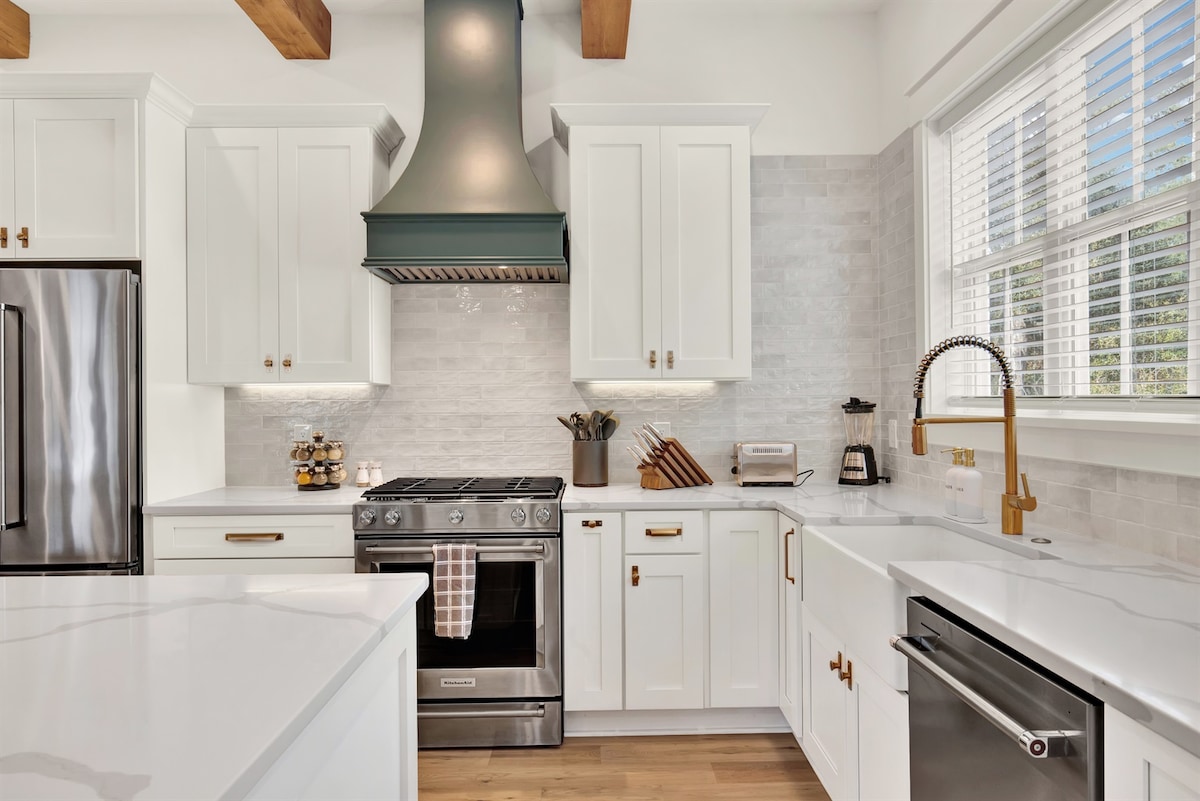 A modern kitchen features white cabinetry with gold hardware and quartz countertops. A stainless steel gas stove is complemented by a large green hood, while natural light floods in through a row of windows. Cookware and utensils are neatly arranged, suggesting an organized space.