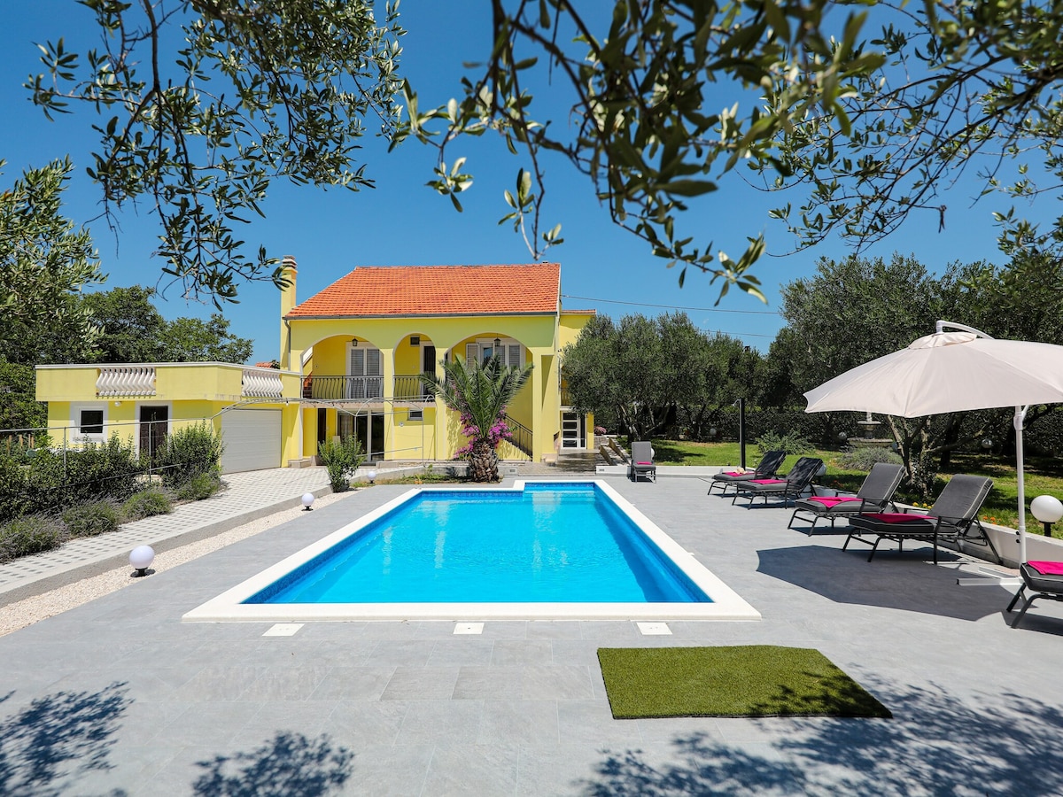 A bright yellow villa is set against a clear blue sky, featuring a vibrant pool surrounded by loungers. Olive trees provide shade, and colorful flowers add natural beauty to the landscaped surroundings. A large umbrella is positioned near the pool, enhancing outdoor relaxation.