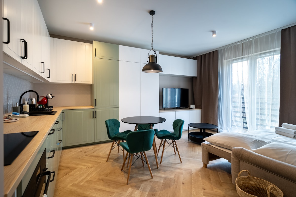 A modern kitchenette equipped with sleek cabinets and essential appliances is visible alongside a round dining table surrounded by four blue chairs. A cozy corner features a sofa, and large windows provide natural light, enhancing the inviting atmosphere of the living area.