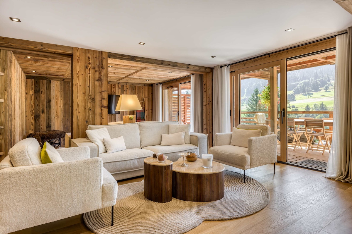 A spacious living area features soft white seating arranged around a round wooden coffee table. Natural light floods the room through large glass doors that open to the balcony, which offers scenic views of the surrounding landscape. Warm wood accents add to the inviting decor.