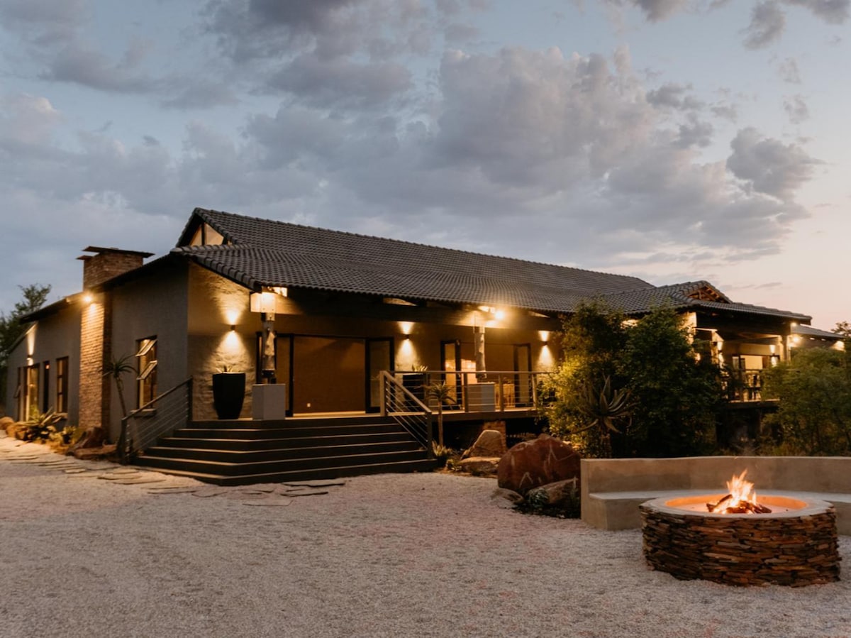 A modern two-story structure is highlighted by outdoor lighting, illuminating the front entrance and inviting seating area. A circular fire pit surrounded by stone complements the gravel landscape, while trees and natural rock features add to the serene atmosphere.