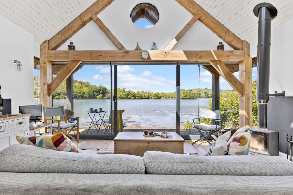 A spacious living area features large sliding doors that open to scenic waterside views. A soft gray sofa is centered in the room, complemented by a wooden coffee table. Natural light fills the area, highlighting a cozy log burner and decorative elements on the mantel.
