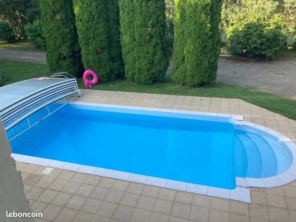 The shared pool is presented, surrounded by tall green hedges that provide privacy. The pool features a light blue water surface and is equipped with a cover. The tiled area surrounding the pool offers easy access and space for lounging.