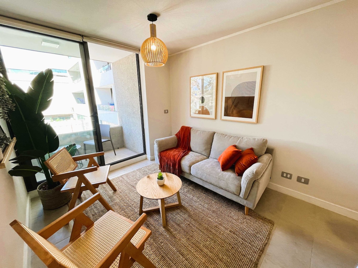 A modern living area features a light grey sofa adorned with orange cushions. Two wooden chairs are positioned nearby, and a small round table sits in the center. Natural light enters through expansive glass doors leading to a balcony, enhancing the room's open and airy feel.