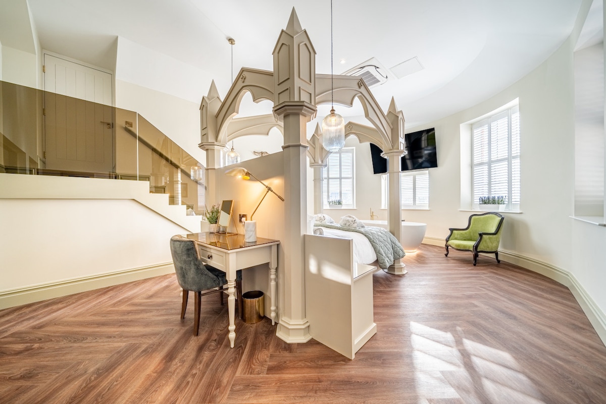 An elegantly designed room features a four-poster bed with a delicate framework. A writing desk and chair are positioned nearby, while large windows allow natural light to fill the space. A comfortable armchair is visible in the corner, enhancing the cozy atmosphere.