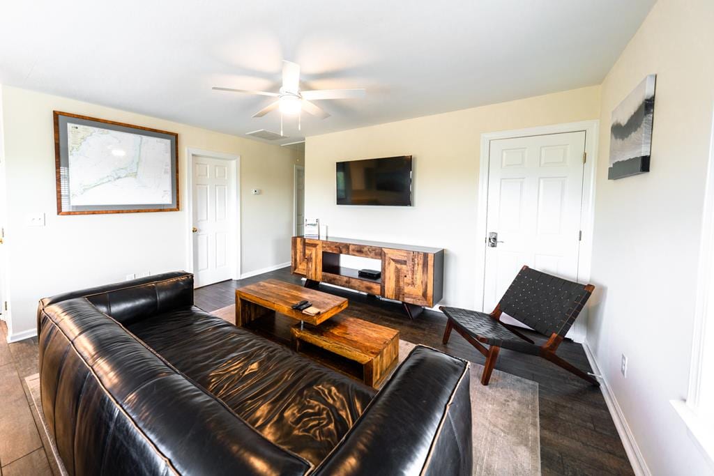 A living room features leather furniture arranged for comfort. A wooden coffee table centers the space, while a wall-mounted smart TV is positioned above a media console. Natural light from a nearby window brightens the room, complemented by a ceiling fan.